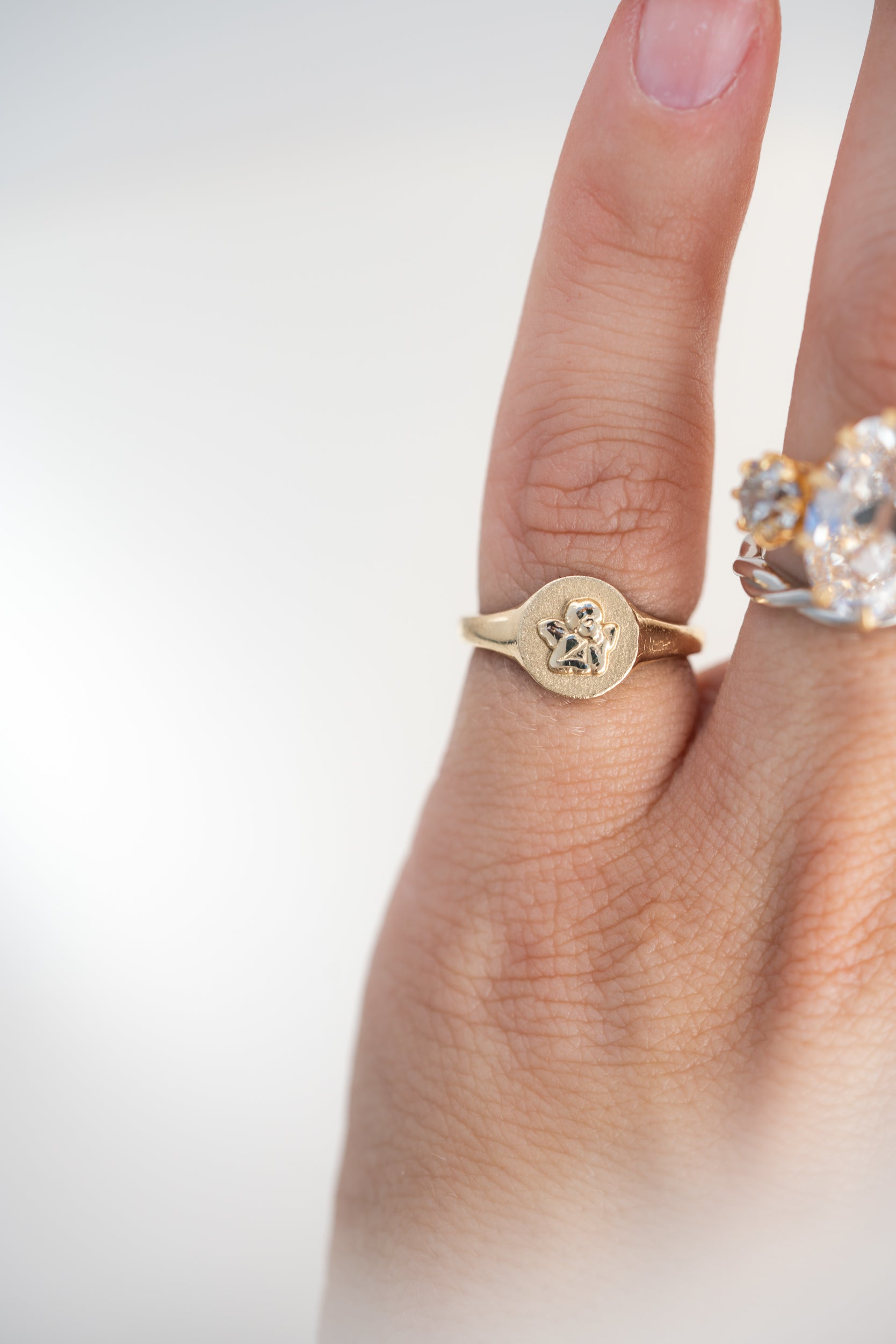 A gold signet ring with a circular flat face and a small cherub shown worn on a person's finger against a soft light gray background.