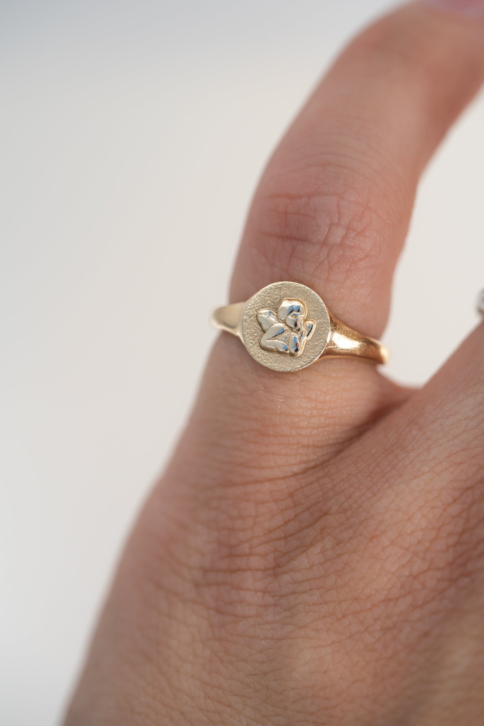 A gold signet ring with a circular flat face and a small cherub shown worn on a person's finger against a soft light gray background.