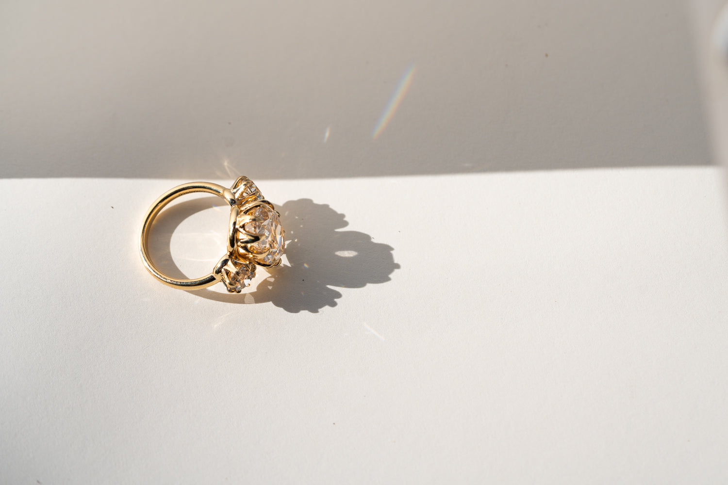 A gold ring with a large clear gemstone set in an ornate, textured setting, resting on a white surface with a hard shadow and a small rainbow reflection under bright sunlight.