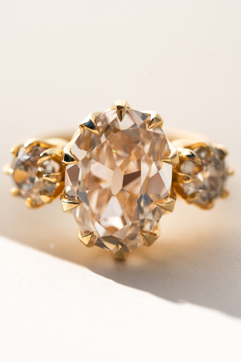 A gold ring featuring a large oval pale pinkish gemstone in a prong setting, flanked by two smaller round clear stones on a warm off-white background.
