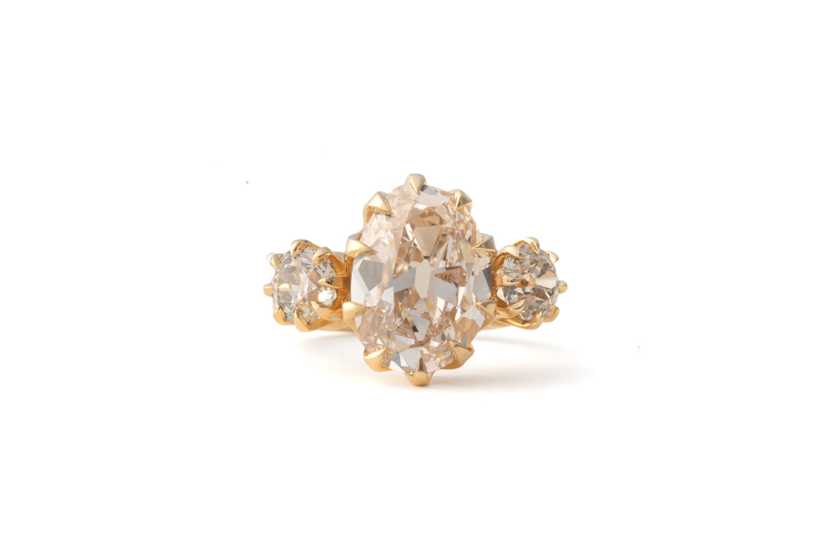 A vintage-style gold ring featuring a large oval pale pinkish gemstone in the center, flanked by two smaller round clear stones on a white background.