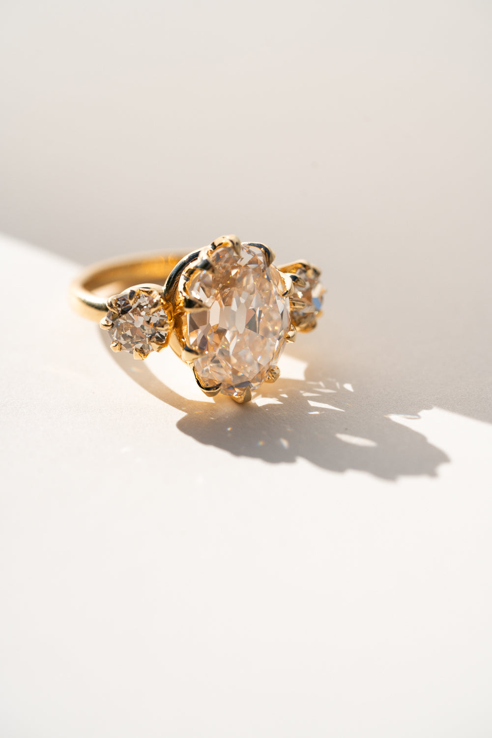 A gold ring featuring a large oval pale pinkish gemstone in the center, flanked by two smaller round stones on the band, photographed on a white surface with strong sunlight and shadows.
