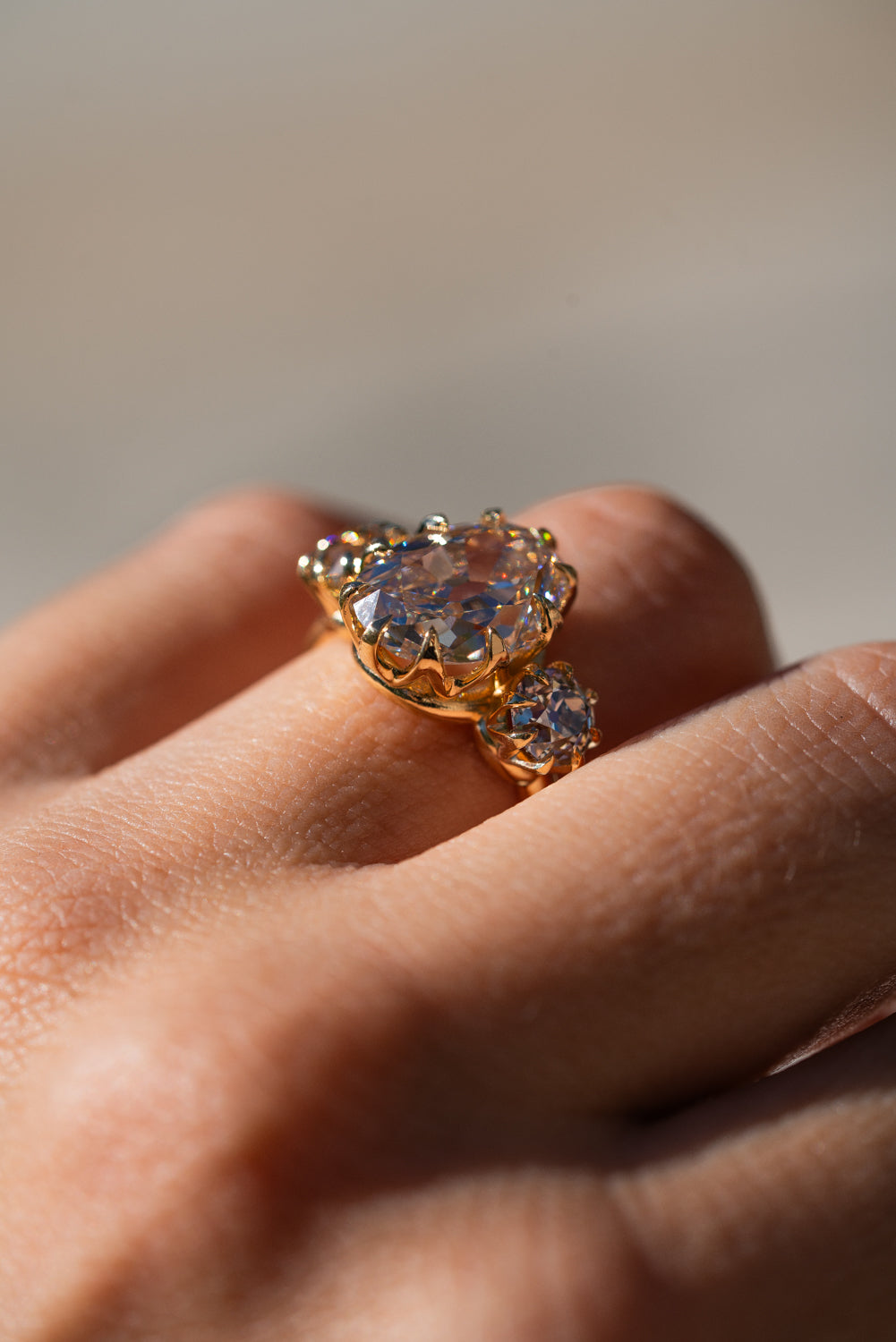 A gold ring with a large oval clear gemstone and a smaller round gemstone, worn on a person's finger against a soft beige background.