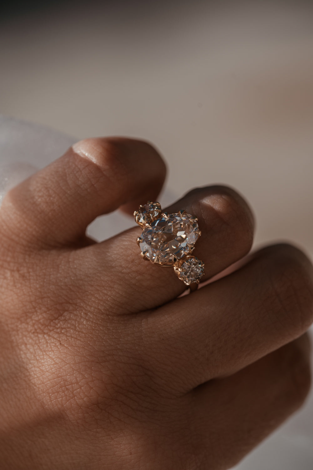A close-up of a hand wearing a rose gold ring with a cluster of clear gemstones, set against a soft, out-of-focus neutral background.