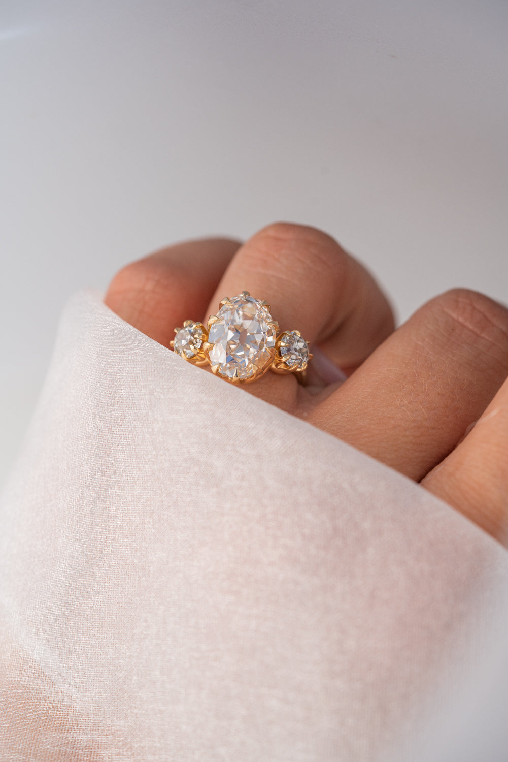 A gold ring with a large oval clear gemstone and smaller side stones, worn on a finger wrapped in a sheer white fabric sleeve.
