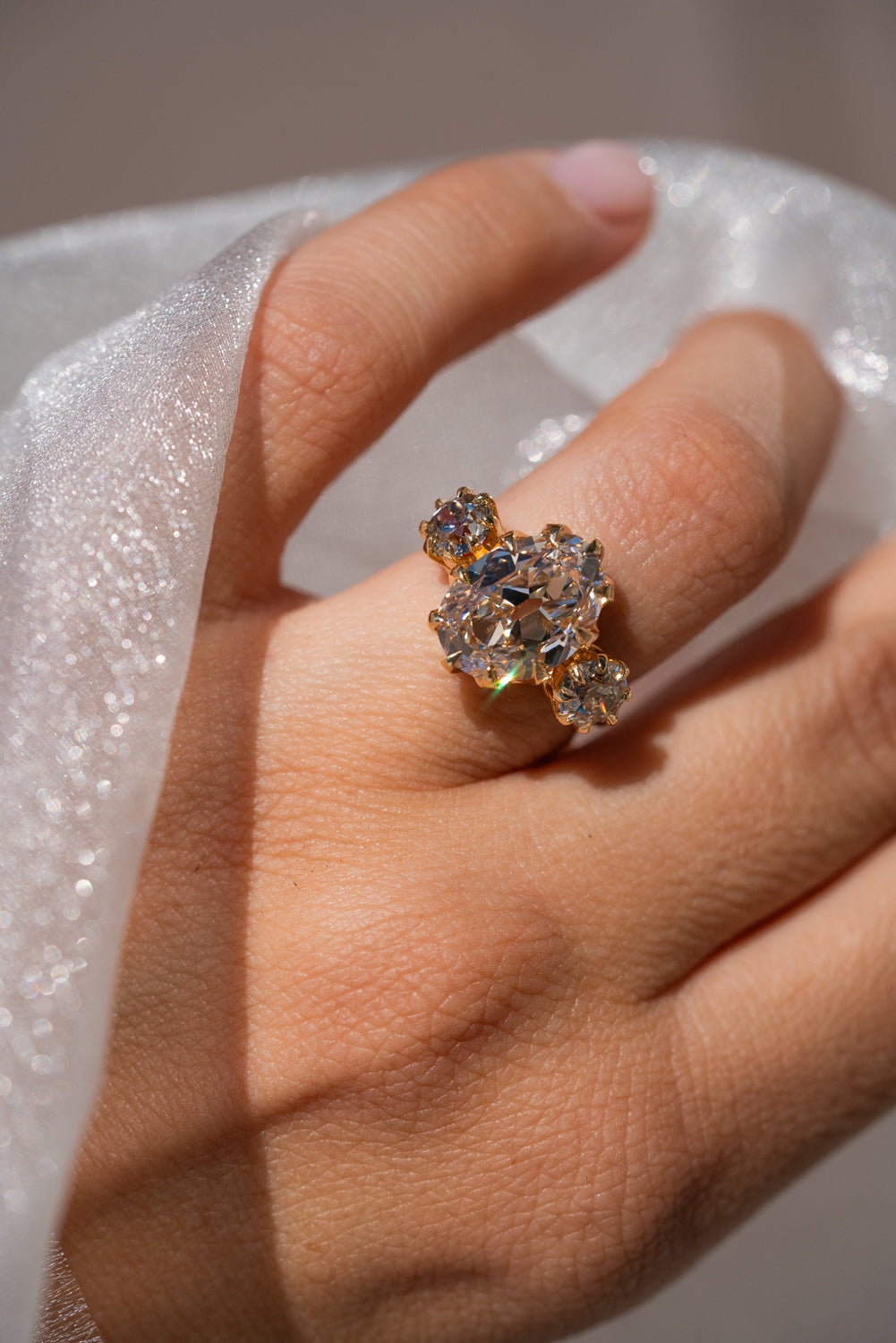A close-up of a hand wearing a gold ring with a cluster of clear gemstones, set against a soft white textured fabric background.